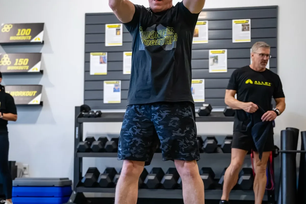 An older member slams battle ropes mid-swing beneath the Stronger Together wall graphic at Alloy Personal Training Royal Oak in Royal Oak, MI.