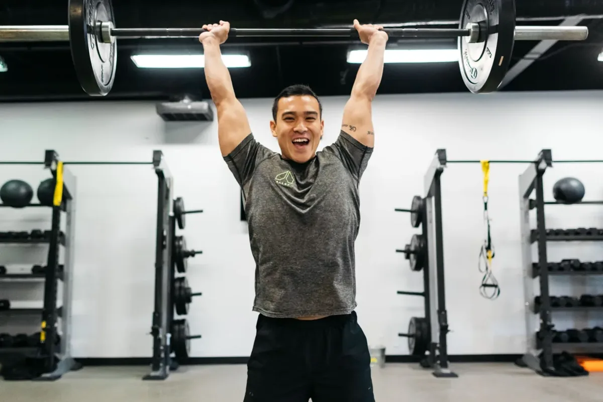 A smiling member locks out a barbell overhead on the Alloy floor logo at Alloy Personal Training Royal Oak in Royal Oak, MI.