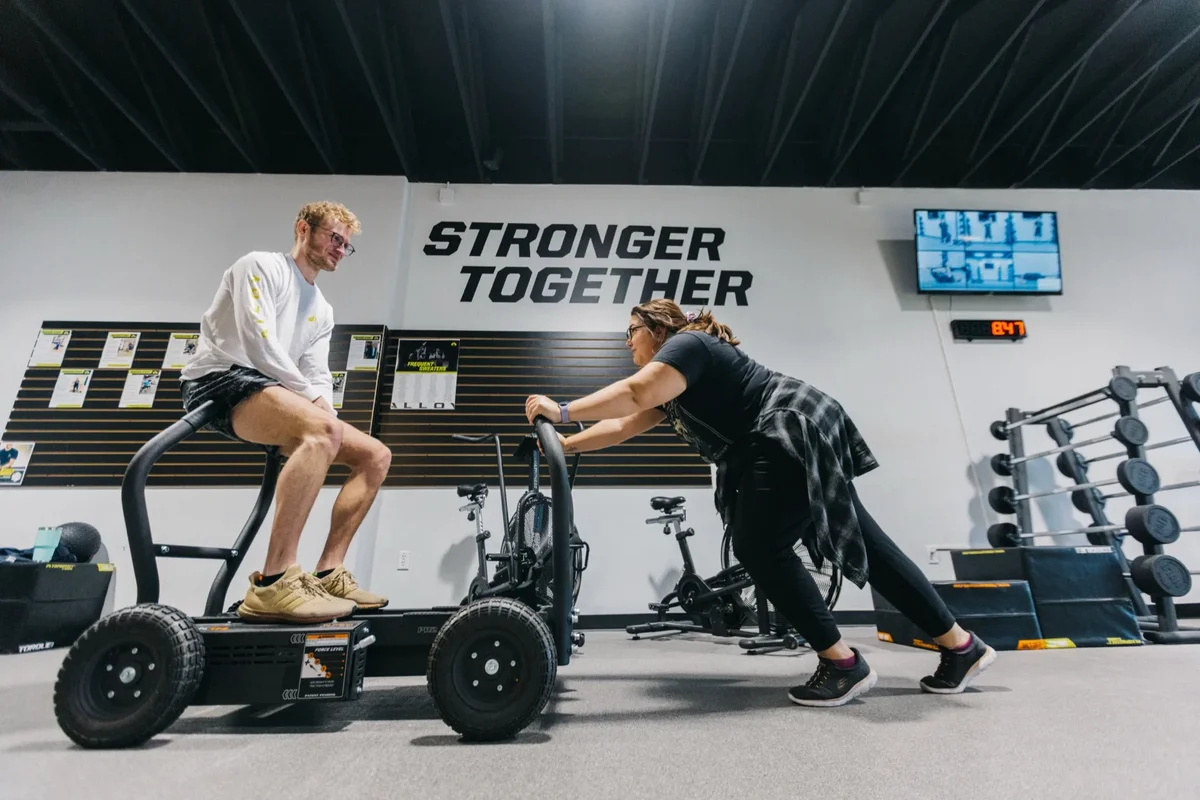 Two members work together on a weighted sled push across the training floor at an Alloy Personal Training studio