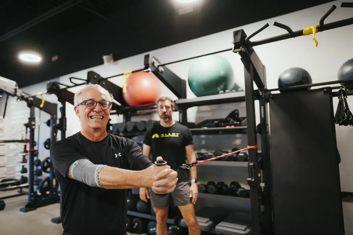 An older member works through resistance band pulls with a trainer nearby at an Alloy Personal Training studio