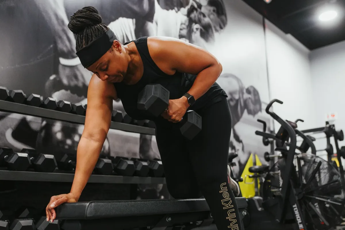 A member performs a single-arm dumbbell row braced on a bench by the dumbbell rack at Alloy Personal Training Royal Oak in Royal Oak, MI.