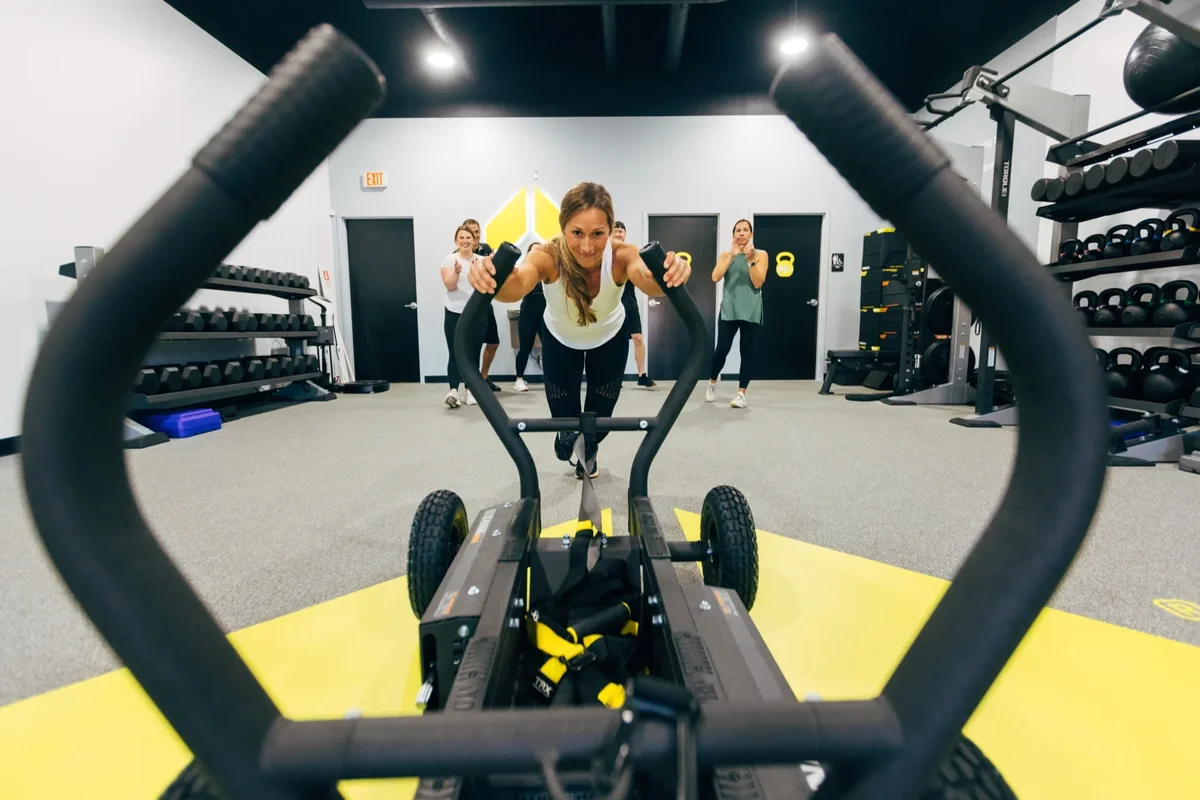 A member pushes a weighted sled toward the camera while others train behind her at an Alloy Personal Training studio